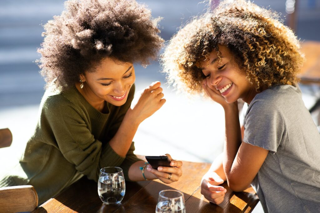 Two happy african female friends at outdoor cafe with cellphone